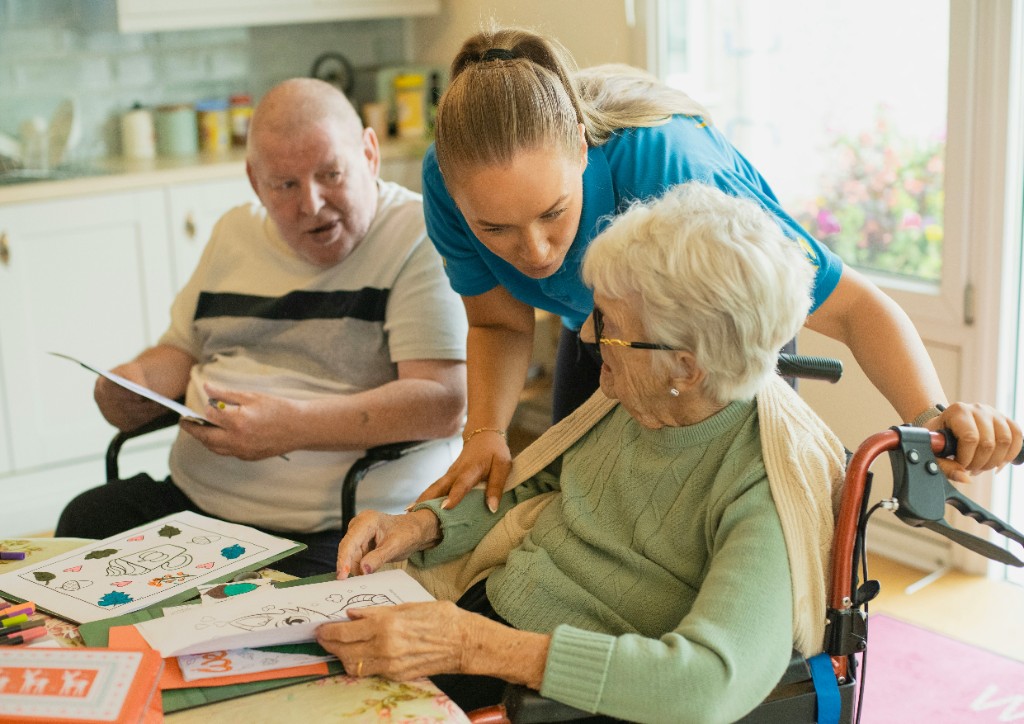 A caregiver helping an older couple with an activity at the kitchen table.